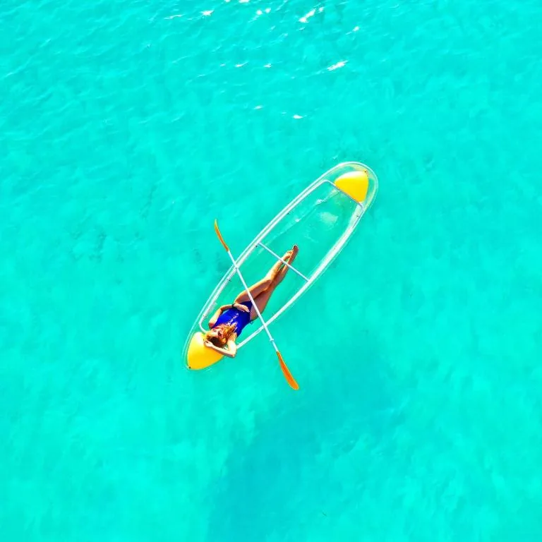 Aerial view of a clear-bottom kayak with yellow paddles in turquoise water, swimmer's legs visible in the hull at Adroit Sunset View, Guraidhoo, Maldives