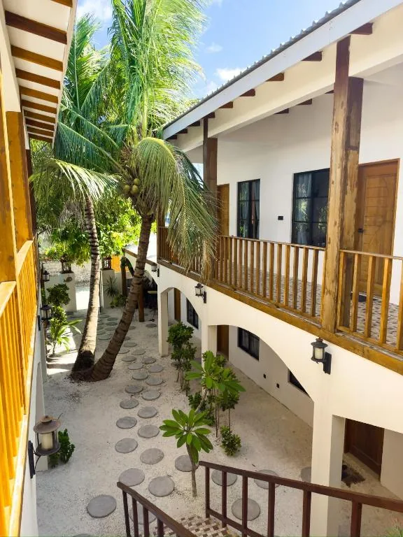 Upper wooden balcony with white railings overlooking a courtyard of stepping-stone path, palm trees and arched ground-floor doors at Ahiva Village, Fulhadhoo, Maldives