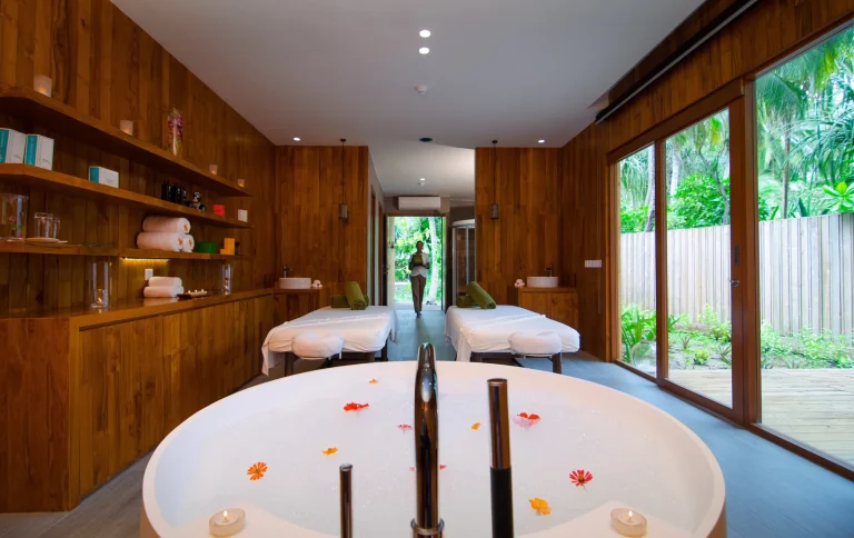 Timber spa treatment room with a petal-filled bath and two massage beds facing the garden at Javvu Spa, Amilla
