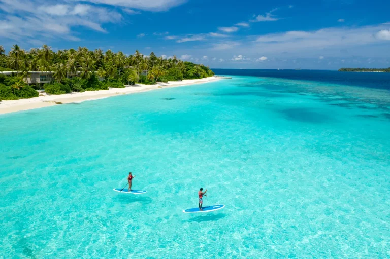 Aerial view of two paddle boarders on a bright turquoise lagoon beside a long white beach at Amilla Maldives
