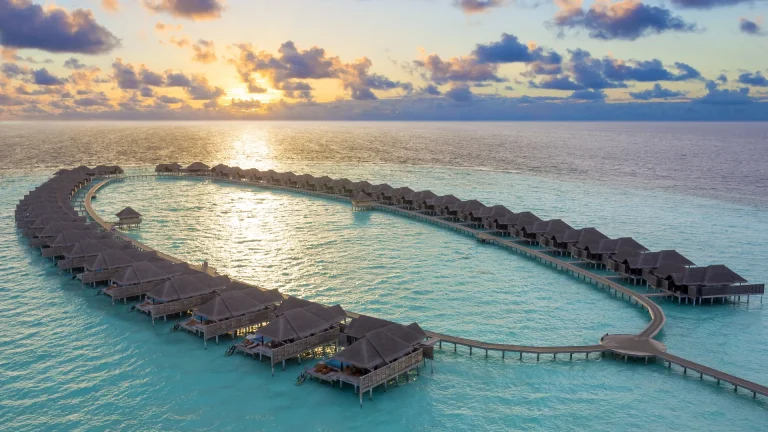 Aerial view of a long curved line of overwater pool villas in a calm lagoon at sunset, Anantara Kihavah, Baa Atoll