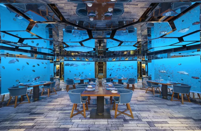 Underwater restaurant with tables set beside large windows full of fish and coral at SEA, Anantara Kihavah