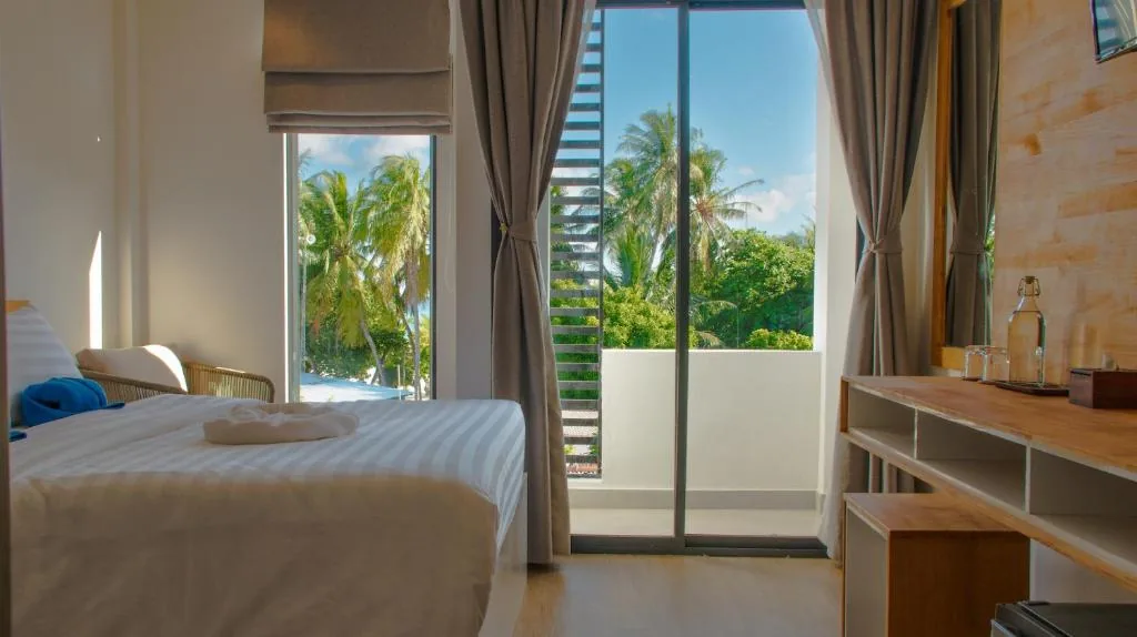 Room with sliding glass door to balcony overlooking palm trees, bed with rolled towels, desk with bottled water and mini-fridge at Atoll Retreat, Fulidhoo, Maldives