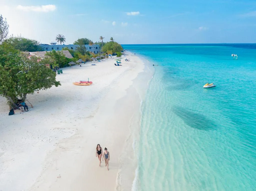 White-sand beach with turquoise water, palm-fringed shore, kayaks and a jet ski, two walkers in foreground at Ayala Ocean View, Gulhi, Maldives