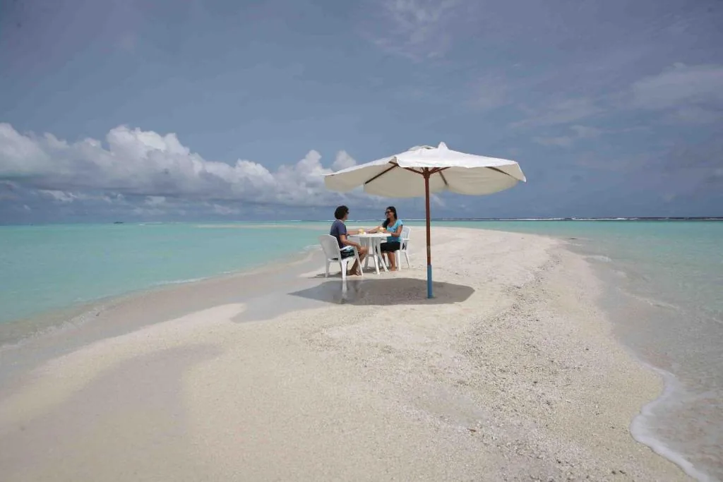 Two people dining on a white-sand beach with a large parasol, turquoise lagoon and distant reef in the background at Azoush Tourist Guesthouse, Fulhadhoo, Maldives