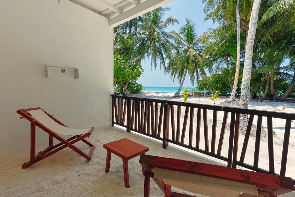 Private balcony with wooden railing, two deck chairs and low table overlooking sandy beach, turquoise sea and palm trees at Azul Retreat, Dhigurah, Maldives