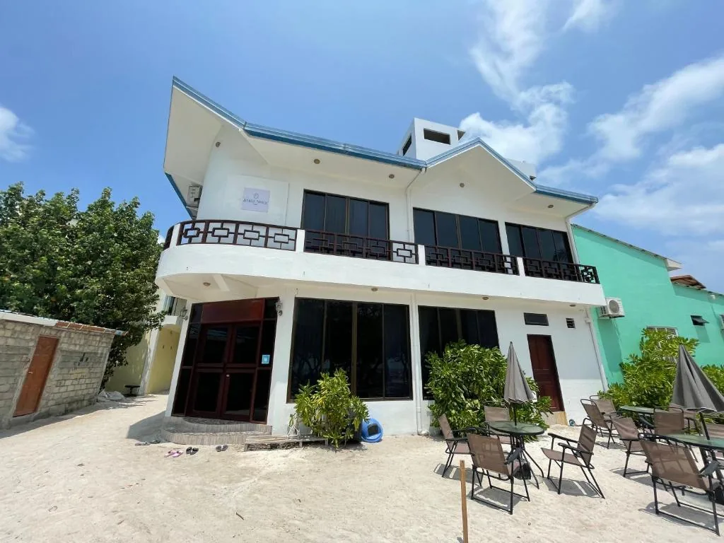 Two-story guesthouse with 'Beach Walk' sign, ground-floor glass doors, tiled roof and outdoor deck with chairs and umbrellas at Beach Walk Villa Maldives, Gulhi, Maldives