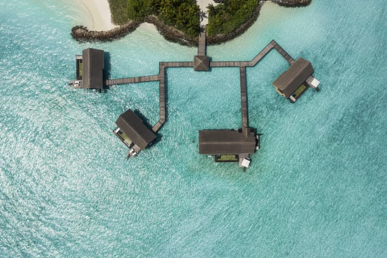 Drone view of the Grand Water Villas at One&Only Reethi Rah, thatched overwater villas joined by a jetty over a shallow turquoise lagoon, North Malé Atoll