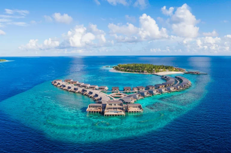 Aerial of The St. Regis Maldives Vommuli Resort, a green island with a curved line of overwater villas and the Iridium Spa on a deep blue Dhaalu Atoll reef