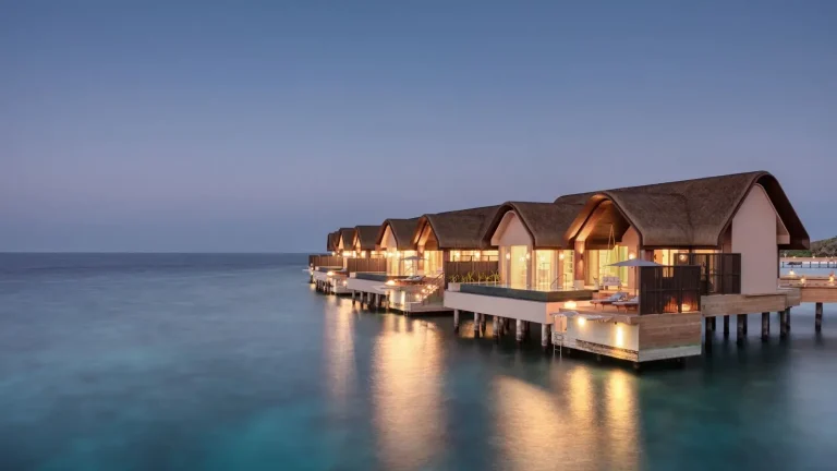 Row of JOALI BEING overwater Ocean Pool Villas lit up at dusk above a calm lagoon, Raa Atoll, Maldives