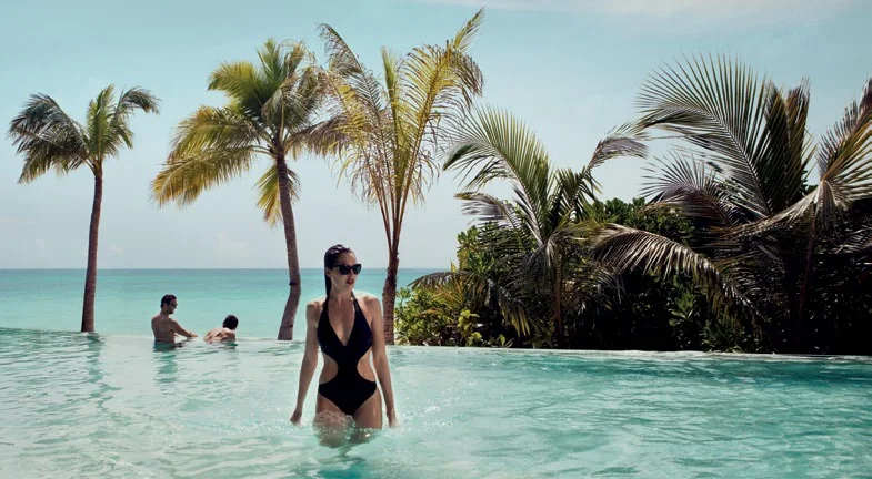 Woman standing in an infinity pool edged by palms with the lagoon beyond, Coco Privé Kuda Hithi, Maldives
