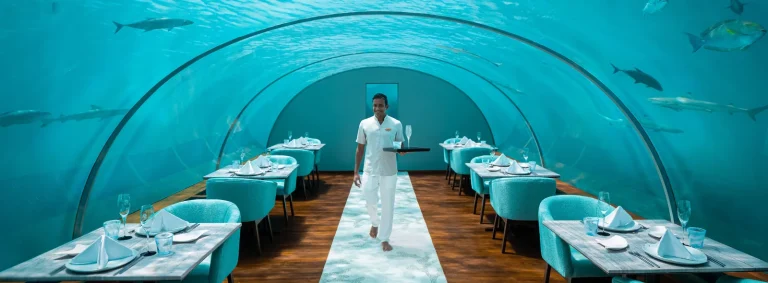 Curved acrylic dining room under the sea with tables set for lunch and a waiter in white, Ithaa Undersea Restaurant, Conrad Maldives Rangali Island