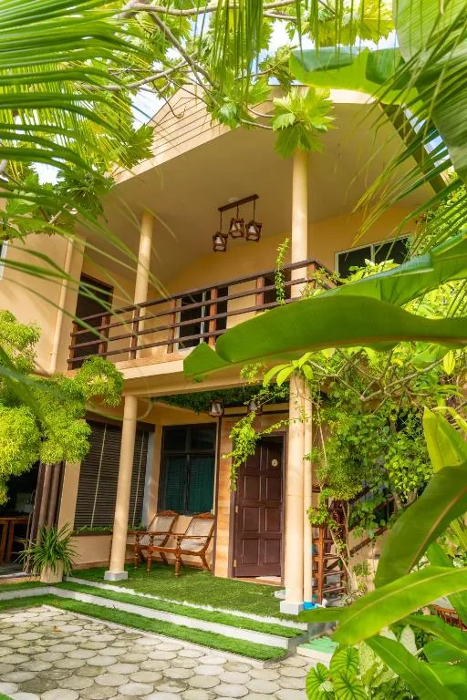 Two-story yellow villa with wooden balcony, hanging lanterns, tropical garden, rattan chairs and dark front door at Daisy Cottage Dhangethi, Dhangethi, Maldives