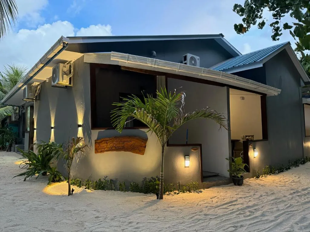 Single-story beachfront bungalow 'Batu Beach Hut' with covered porch, wall lanterns, AC unit and palm tree on sandy path at Dhigurah Beach Inn, Dhigurah, Maldives