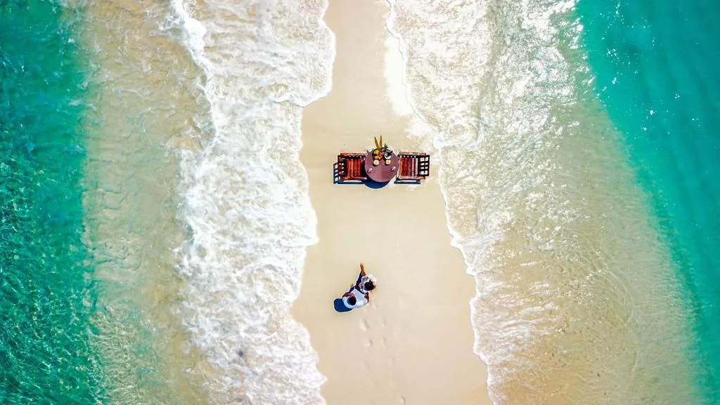 Aerial view of a white-sand beach with turquoise water, a floating platform with a hammock and two people in blue life jackets at Escapism Maldives, Feridhoo, Maldives