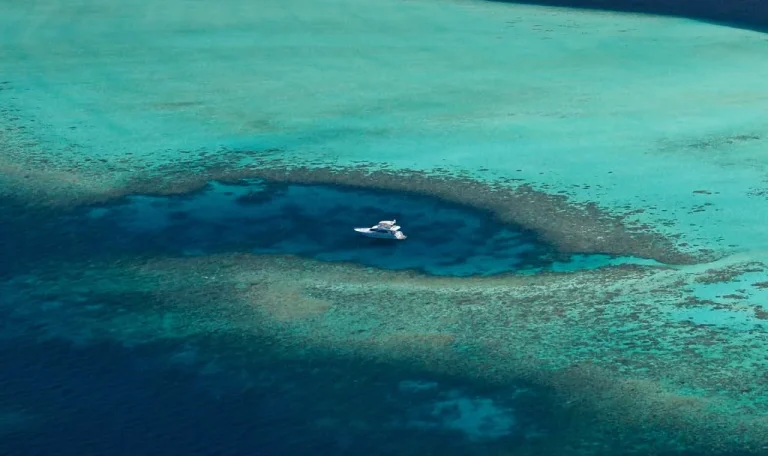 Aerial view of a white yacht anchored in a deep blue reef pool surrounded by turquoise shallows, Baa Atoll, Maldives