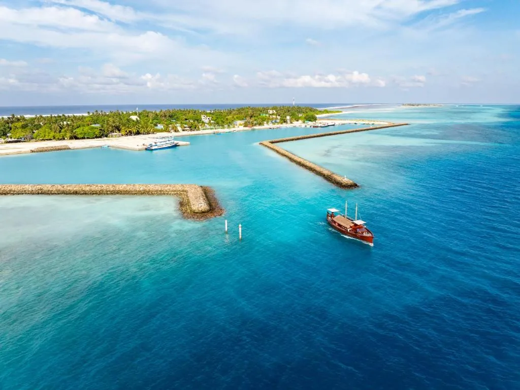 Wooden dhoni passing a coral reef breakwater with a palm-fringed island and turquoise lagoon in the Maldives at Fushi Beach, Fenfushi, Maldives