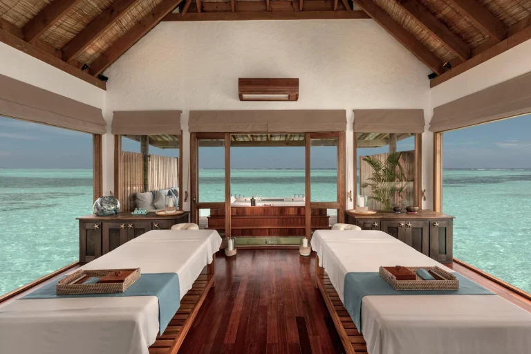Overwater spa treatment room with two massage beds and open doors to the lagoon at Meera Spa, Gili Lankanfushi