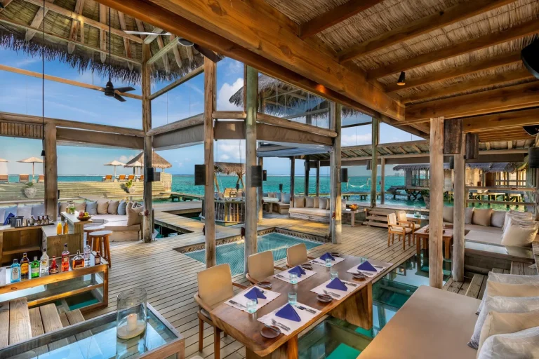 Timber dining room with glass floor panels and tables set over a turquoise lagoon at Gili Lankanfushi