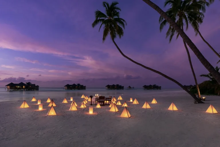 Private dinner table on the sand lit by glowing lanterns under palm trees at dusk at Gili Lankanfushi