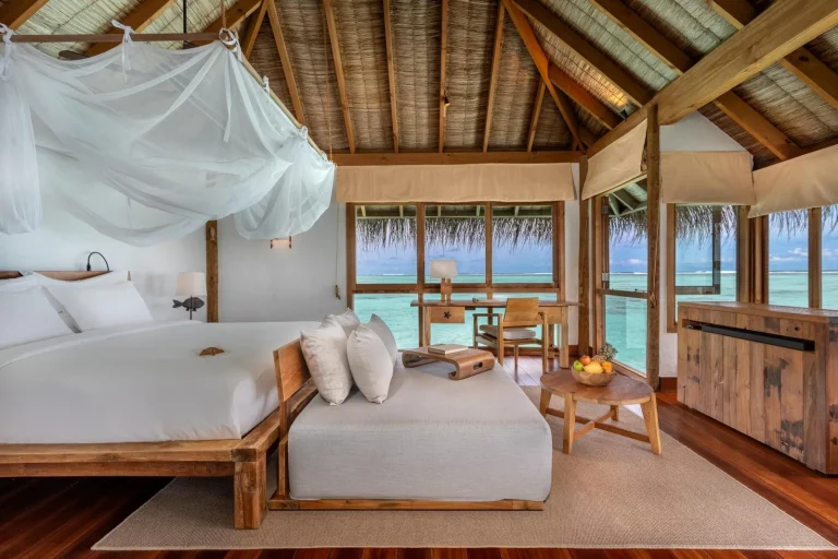 Bedroom with a white canopy bed, timber floors and wide windows over the lagoon in a Gili Lankanfushi villa