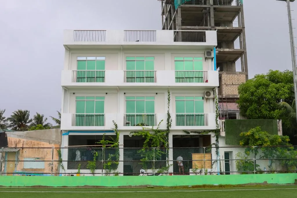 Four-story white building with green-tinted windows, balcony and rooftop terrace beside a green field at Green Vista Maafushi in Maafushi, Maldives