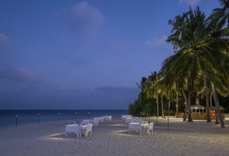 Dinner tables set on the sand beside palms at dusk at The Firepit, The Halcyon Private Isles Maldives
