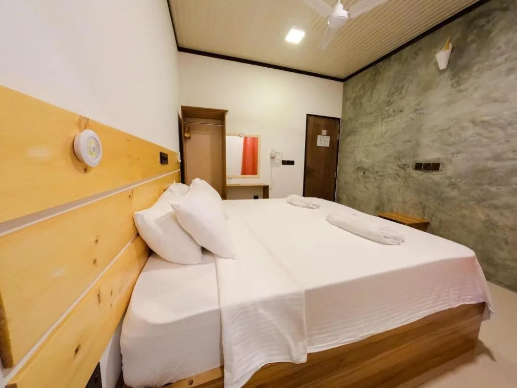 Double bed with wooden headboard, wall-mounted AC and fan, mirror, exposed concrete wall and folded towels on bed at Huvan Inn, Fulidhoo, Maldives