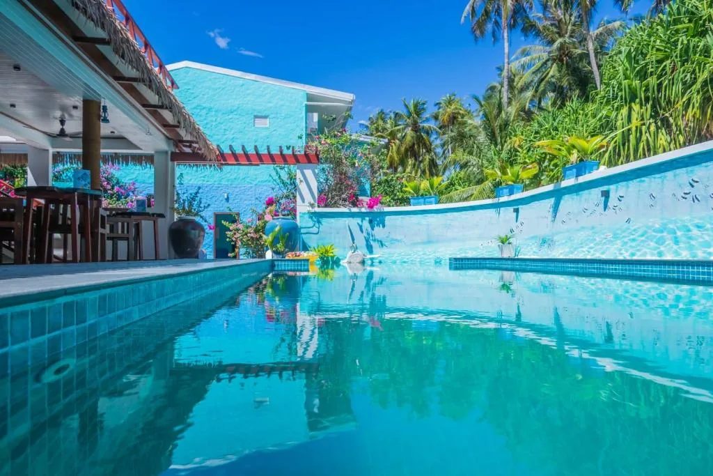 Outdoor pool with tiled deck, shaded lounge, tropical plants and palm-tree wall at a blue-tiled guesthouse at Island Luxury Dive, Fulhadhoo, Maldives