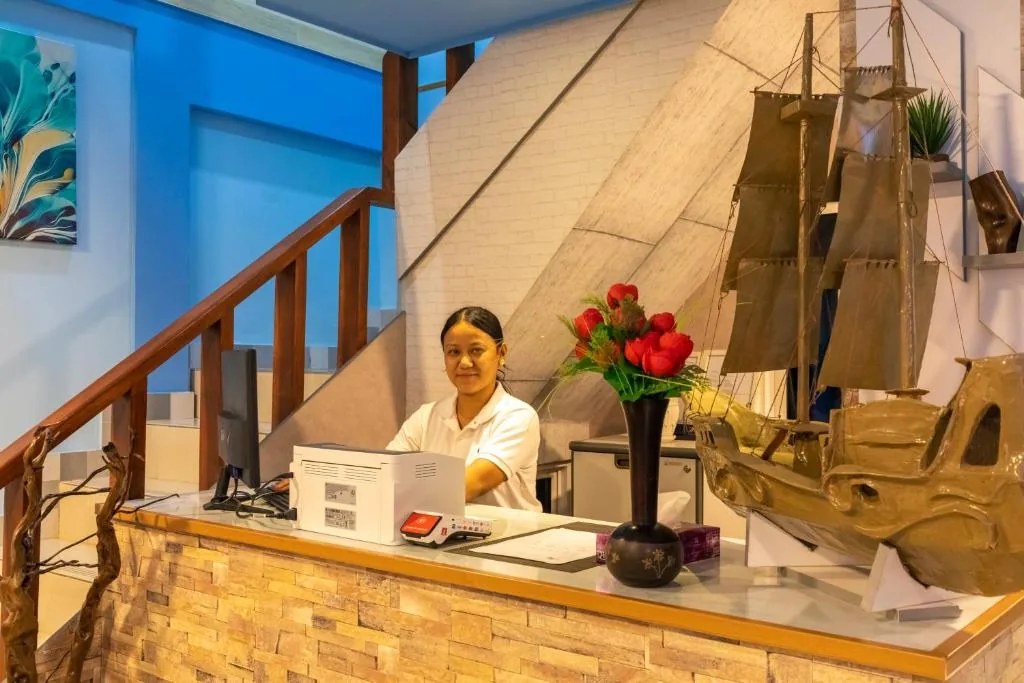 Reception desk with stone counter, computer, card reader and vase of red flowers, sailboat decor and staff at front desk at Isle Inn, Fuvahmulah, Maldives