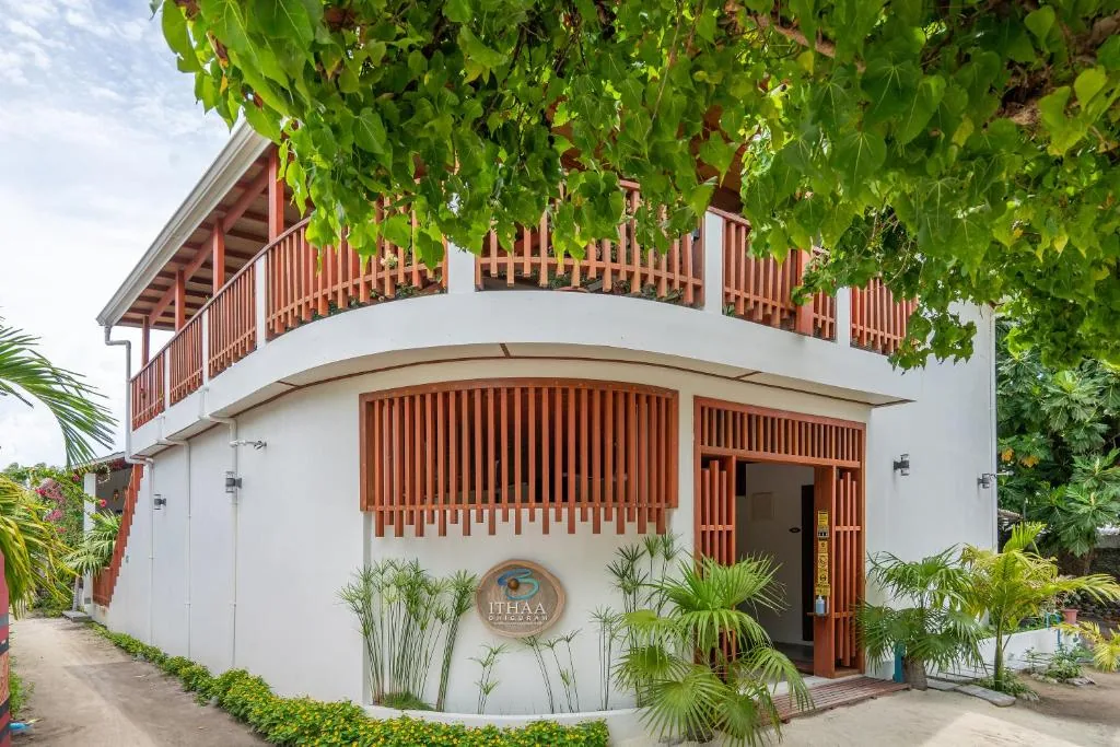 Two-story guesthouse with wooden balcony, tropical plants and green 'ITHAA SIDAWA' sign on a sandy path at Ithaa Dhigurah, Dhigurah, Maldives