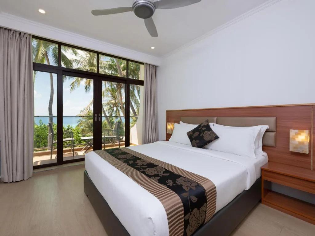 King bed with floral runner, ceiling fan and sliding glass doors to a private balcony with ocean and palm views at Kaani Beach Hotel in Maafushi, Maldives
