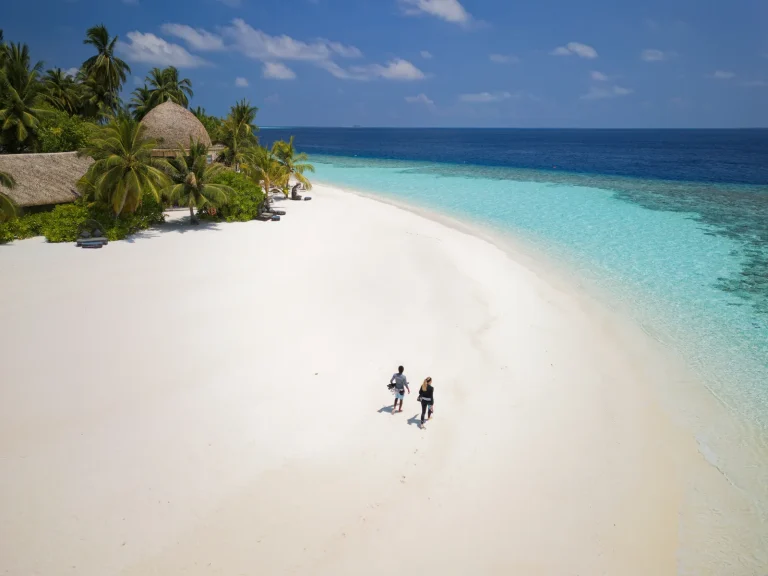A couple walking along a long curve of white sand with palms and thatched villas behind, Kandolhu Maldives, North Ari Atoll