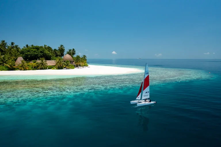 Small sailing catamaran with a red and white sail on turquoise water off the palm-covered island of Kandolhu, Maldives