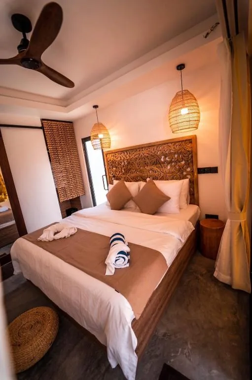 King bed with carved wooden headboard, woven pendant lights, ceiling fan, sliding door and rolled towels on bed at Kunaa Beach Inn, Fulidhoo, Maldives