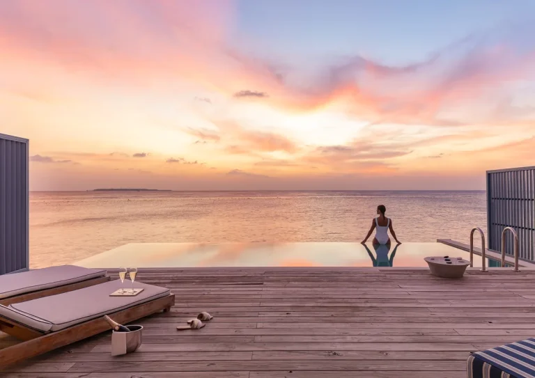 Guest sitting at the end of a private infinity pool on the deck of an Amilla Sunset Water Pool Villa as the sky turns orange, Baa Atoll, Maldives