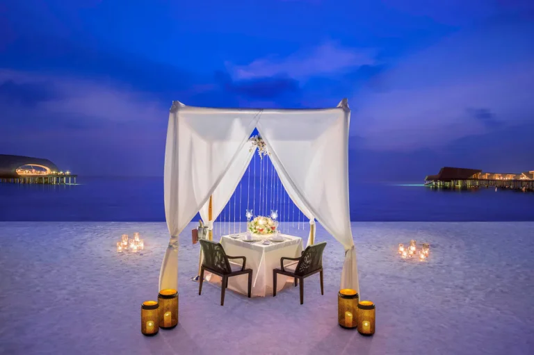 Candlelit dinner table under a white canopy on the beach at dusk with overwater villas in the background at The St. Regis Maldives Vommuli Resort, Dhaalu Atoll