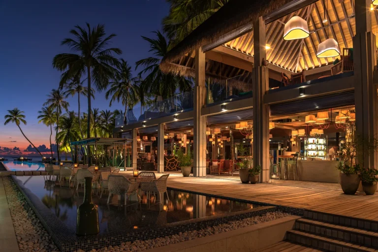 Avi Pool Bar at Velaa Private Island lit up at night, with warm lanterns, a long pool and palm trees at dusk, Noonu Atoll