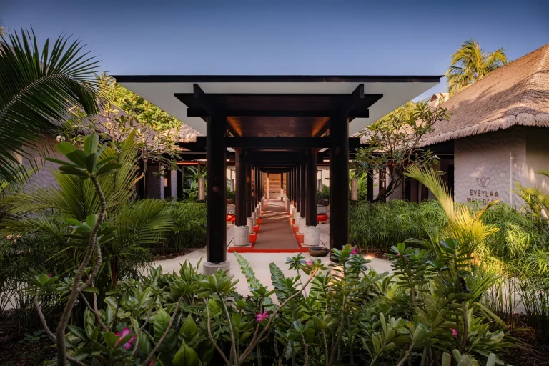 Entrance walkway to the Wellbeing Village at Velaa Private Island, a red-lit path between dark timber pavilions and tropical gardens, Noonu Atoll