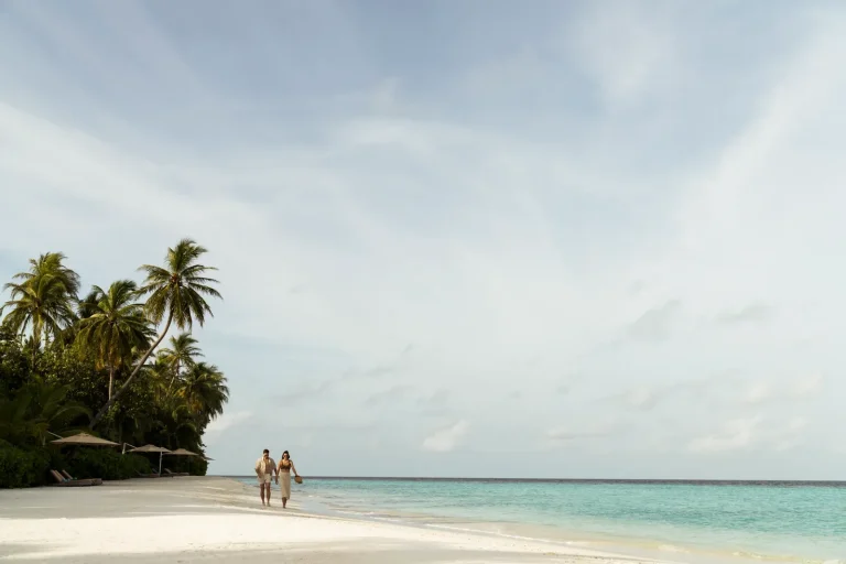 Couple walking along a wide white beach beside a calm turquoise lagoon at Constance Halaveli, North Ari Atoll, Maldives