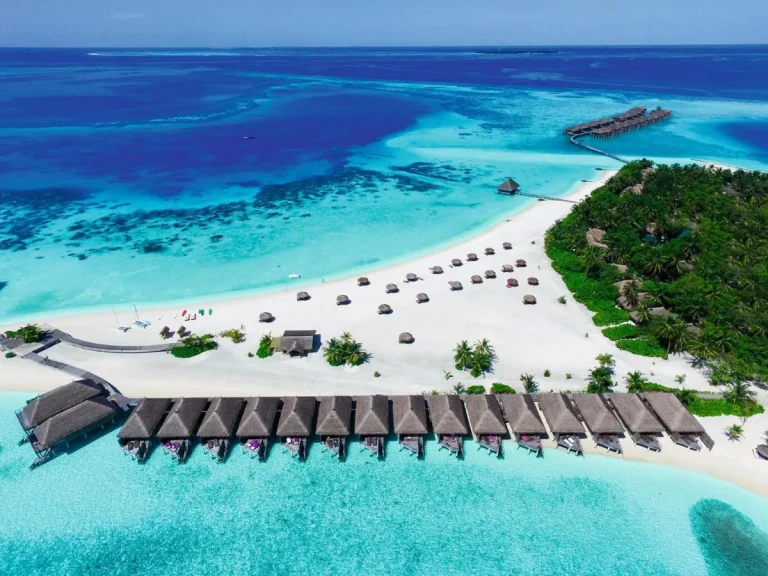 Aerial view of beach villas along white sand and a line of thatched overwater villas on a jetty over a turquoise lagoon at Constance Moofushi