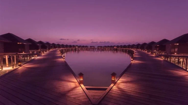 Curved timber jetty lit by lanterns at dusk leading to a line of thatched overwater villas at Lily Beach Resort and Spa, South Ari Atoll