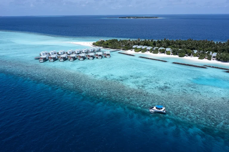 Aerial view of a row of overwater villas on a jetty above a turquoise lagoon and reef edge, with the resort island beyond, Raa Atoll, Maldives