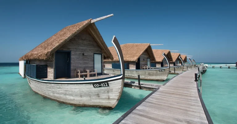 Row of thatched water villas shaped like traditional dhoni boats along a timber jetty over clear water at COMO Cocoa Island, South MalΓ© Atoll, Maldives