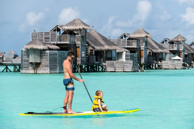 Father and young child in a life jacket paddleboarding past overwater villas at Gili Lankanfushi, North MalΓ© Atoll, Maldives