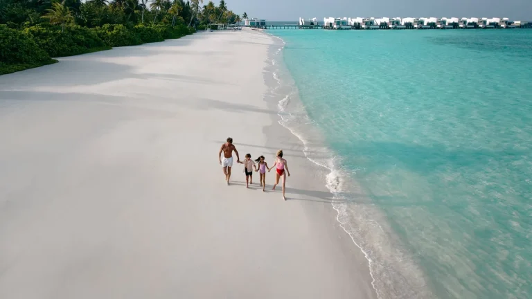 Family of four walking hand in hand along a white sand beach beside a turquoise lagoon at Jumeirah Olhahali Island, North MalΓ© Atoll, Maldives