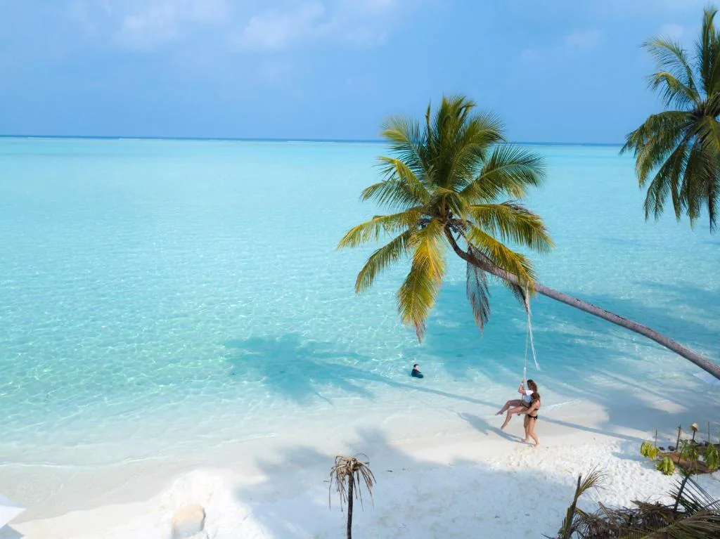 Two people on a rope swing at a palm tree on a white-sand beach with turquoise lagoon and nearby inflatable ring at Masfalhi View Inn, Felidhoo, Maldives