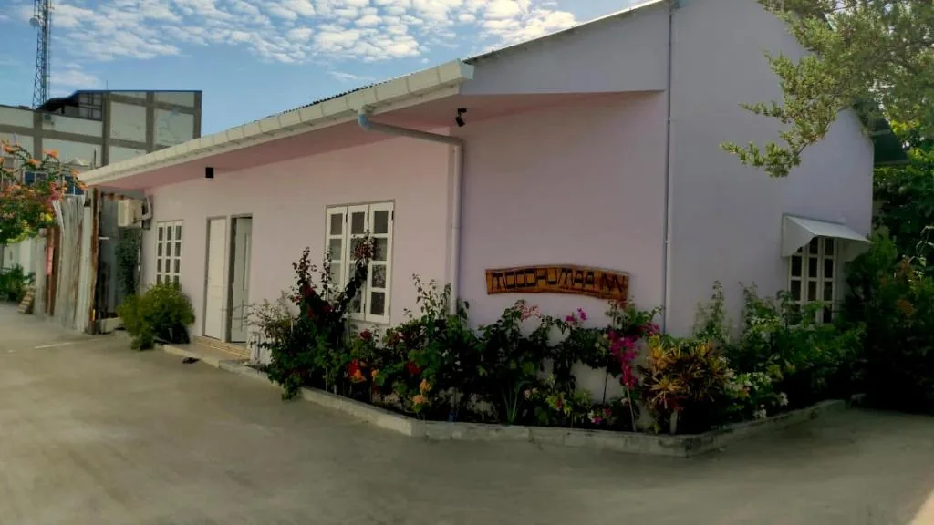 Pink single-story Inn with wooden sign 'MOOD INN', white-framed windows, tropical plants and exterior AC units at Moodhumaa Inn, Guraidhoo, Maldives
