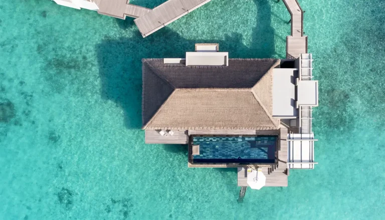 Top-down drone view of a Cheval Blanc Randheli water villa with pitched roof, deck and long private infinity pool over a clear turquoise lagoon, Noonu Atoll