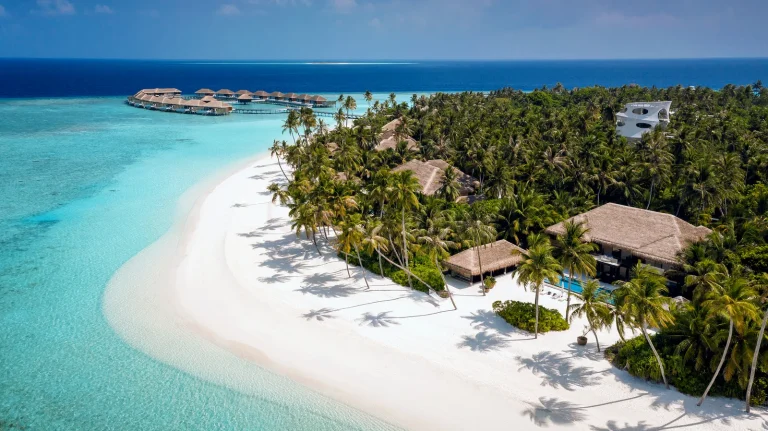 Aerial view of a four-bedroom Velaa Private Residence among palms on a long white beach at Velaa Private Island, with water villas in the distance, Noonu Atoll
